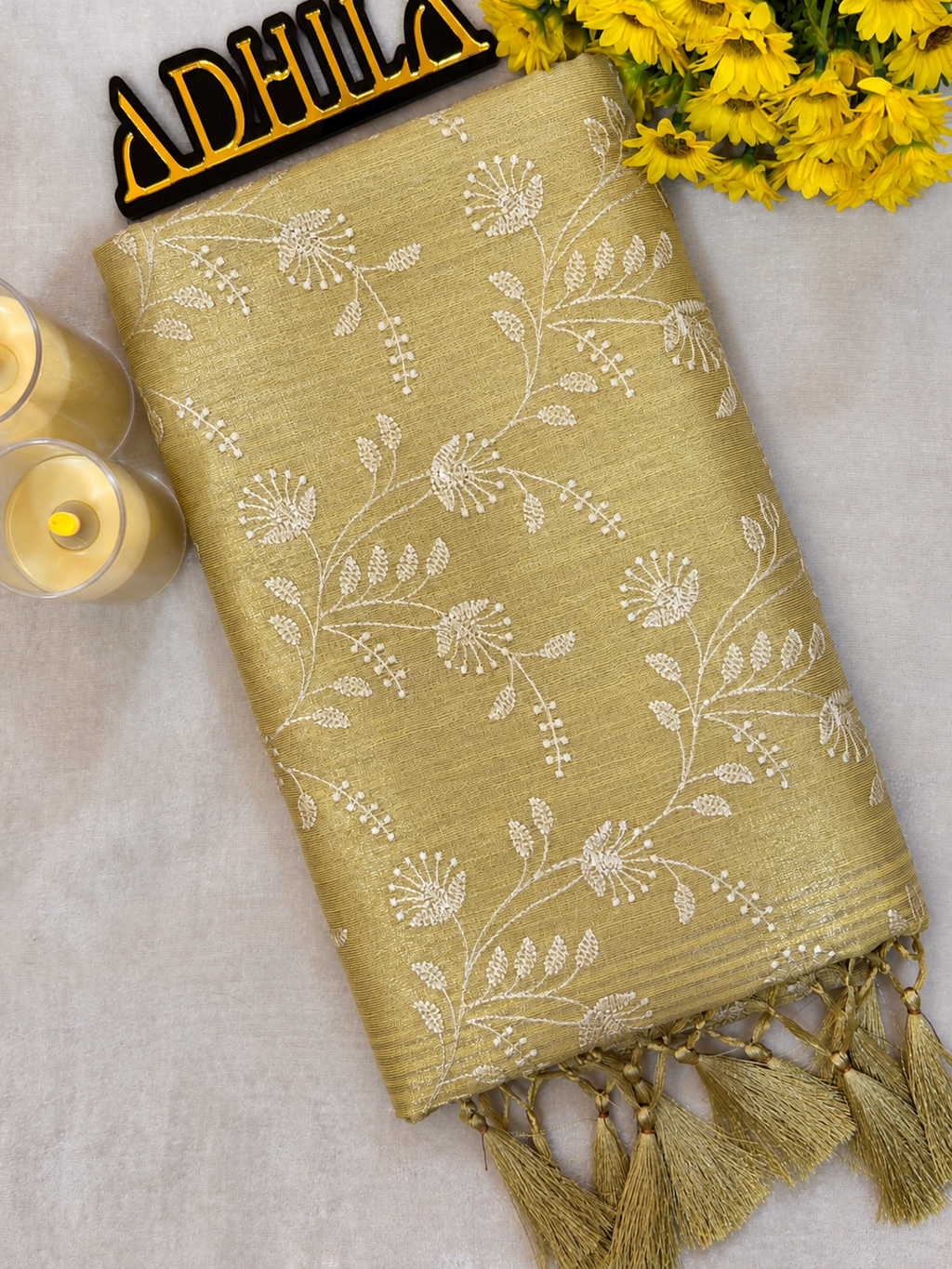 Golden Yellow Floral Tissue Saree – folded view showing floral Jaal work and golden tassels with Adhila logo